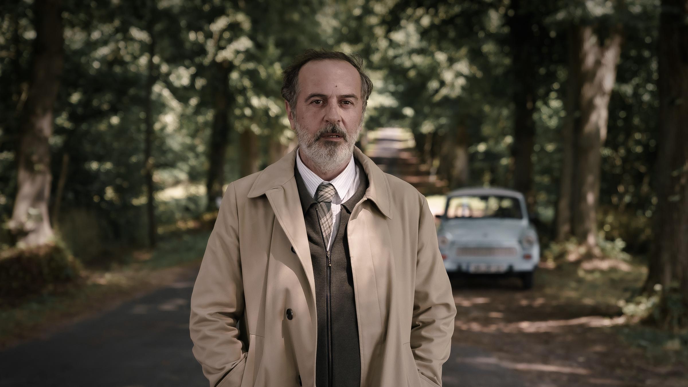 "Doktor Ballouz - Willkommen zurück": Ballouz (Merab Ninidze) steht auf einer Allee, hat die Hände in der Tasche seines Mantels und guckt nach vorne zum Wegesrand. Im Hintergrund ist sein hellblauer Trabbi zu sehen.