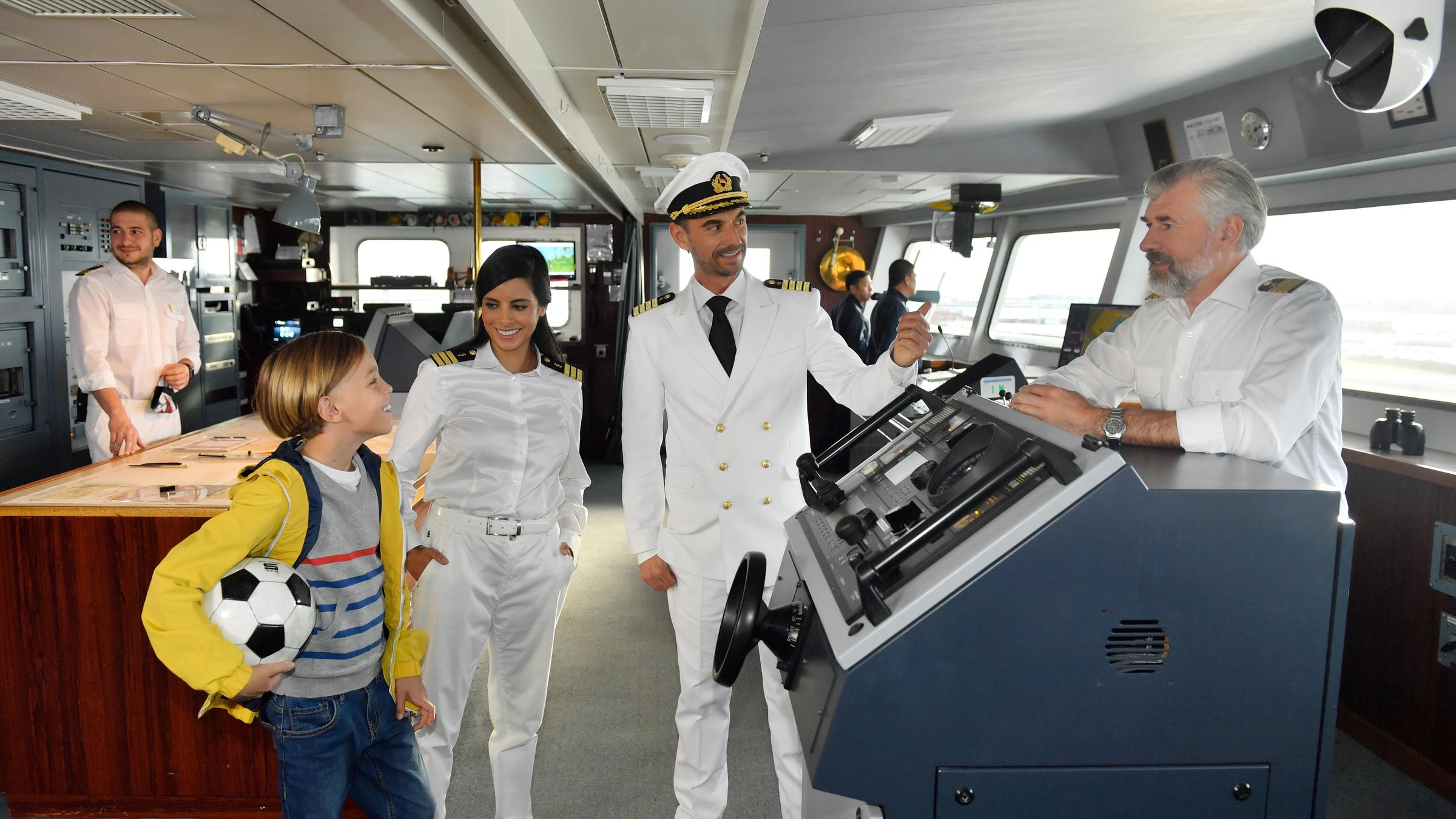 "Das Traumschiff: Malediven/Thaa Atoll": Neben Kapitän Max Parger (Florian Silbereisen) stehen Dr. Delgado (Collien Ulmen-Fernandes) und Timo (Otis Whigham) auf der Kommandobrücke des Schiffs. Kapitän Parger zeigt mit dem Finger auf Staffkapitän Martin Grimm (Daniel Morgenroth), der am Steuerstand lehnt. Timo hält einen Fußball unterm Arm.
