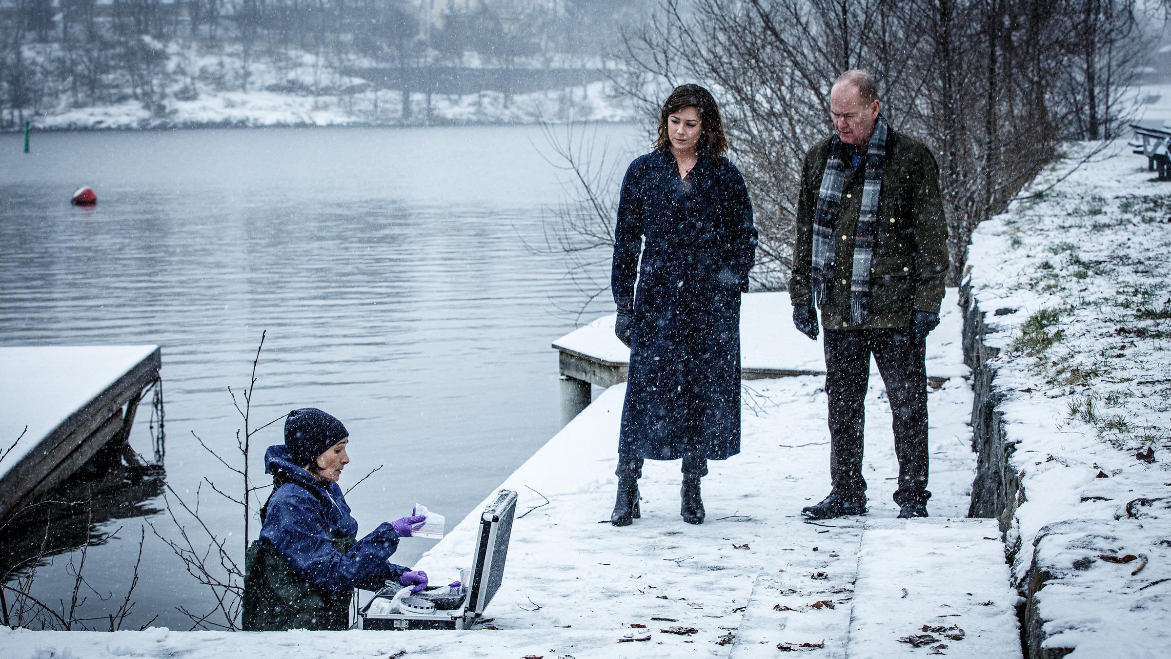 "Kommissar Beck: Undercover": An einem verschneiten Flussufer stehen Alexandra Beijer (Jennie Silfverhjelm) und Martin Beck (Peter Haber) und sprechen mit der Pathologin Bergström (Åsa Karlin). Diese steht im Wasser und hat einen Equipment-Koffer vor sich aufgebaut.