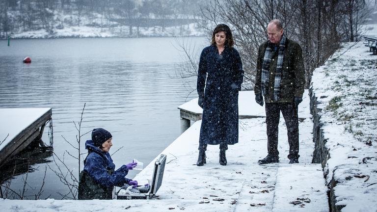 "Kommissar Beck: Undercover": An einem verschneiten Flussufer stehen Alexandra Beijer (Jennie Silfverhjelm) und Martin Beck (Peter Haber) und sprechen mit der Pathologin Bergström (Åsa Karlin). Diese steht im Wasser und hat einen Equipment-Koffer vor sich aufgebaut.