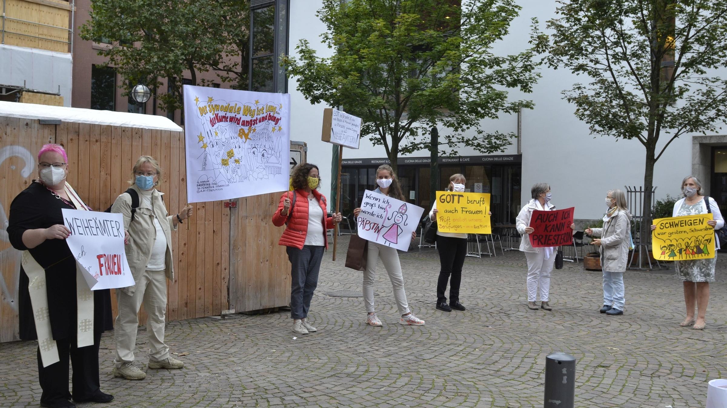 "Frau. Macht. Kirche. - Eine Institution gerät ins Wanken": Eine Gruppe von Frauen steht auf einem Platz und hält ihre bunten Protestschilder in den Händen.