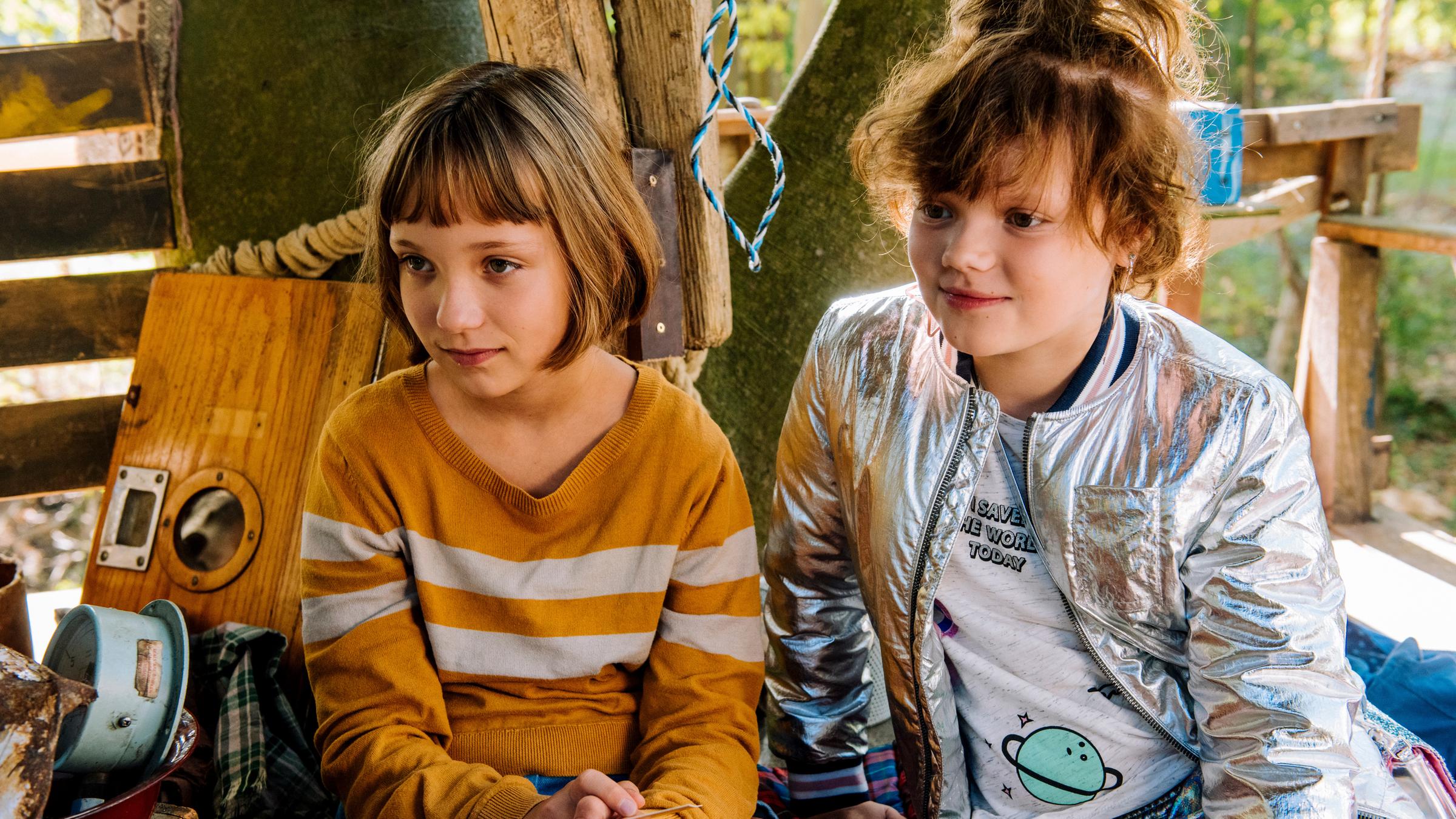 "Mein Lotta-Leben - Alles Bingo mit Flamingo!": Lotta (Meggy Hussong) sitzt neben Cheyenne (Yola Streese) auf dem Boden in ihrem Baumhaus. Lotta hält eine Einladungskarte in den Händen. Die Mädchen lächeln.