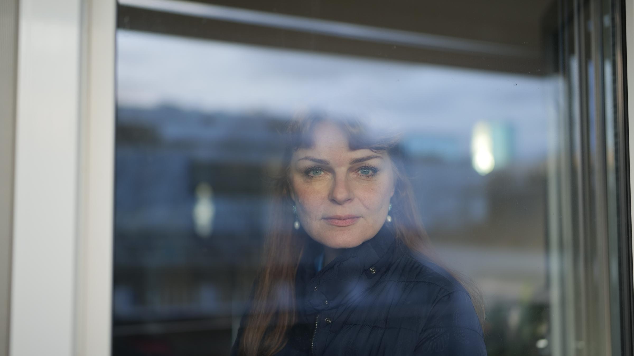 "37°- Wisch und weg ": Portrait junge Frau mit braunen langen Haaren. Sie steht hinter einem Fenster und lächelt in die Kamera.