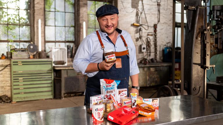 "ZDFzeit - Die Tricks von Ferrero": Sebastian Lege steht in seiner Werkstatt vor einem Tisch voller Produkte von Ferrero und lacht in die Kamera.