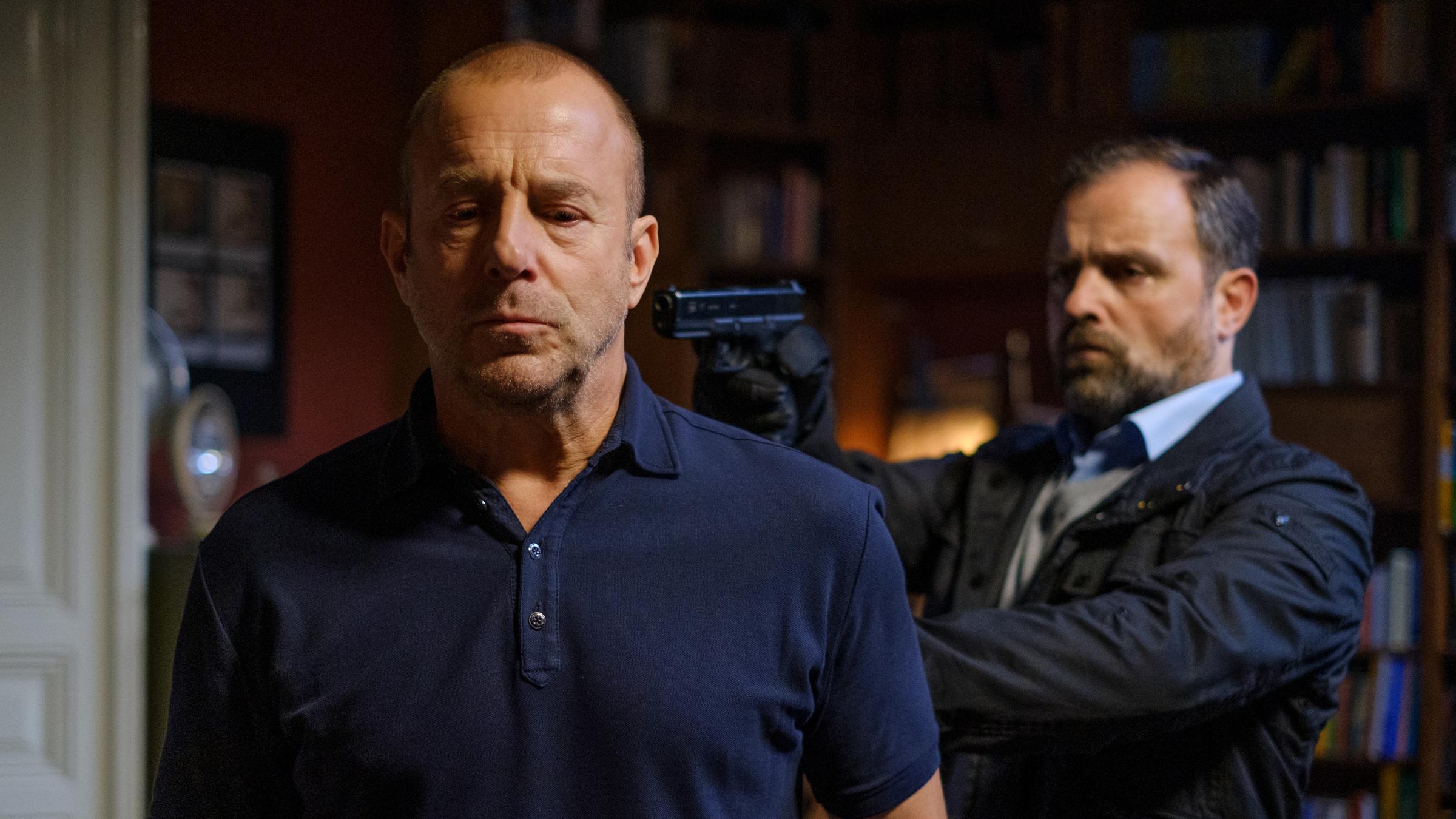 "Spuren des Bösen - Schuld": Gerhard Mesek (Juergen Maurer) steht entschlossen rechts hinter Richard Brock (Heino Ferch) und hält ihm eine Pistole an den Hinterkopf. Richard Brock schaut angespannt zu Boden. Im Hintergrund sieht man lauter hohe Regalschränke voll mit Büchern.