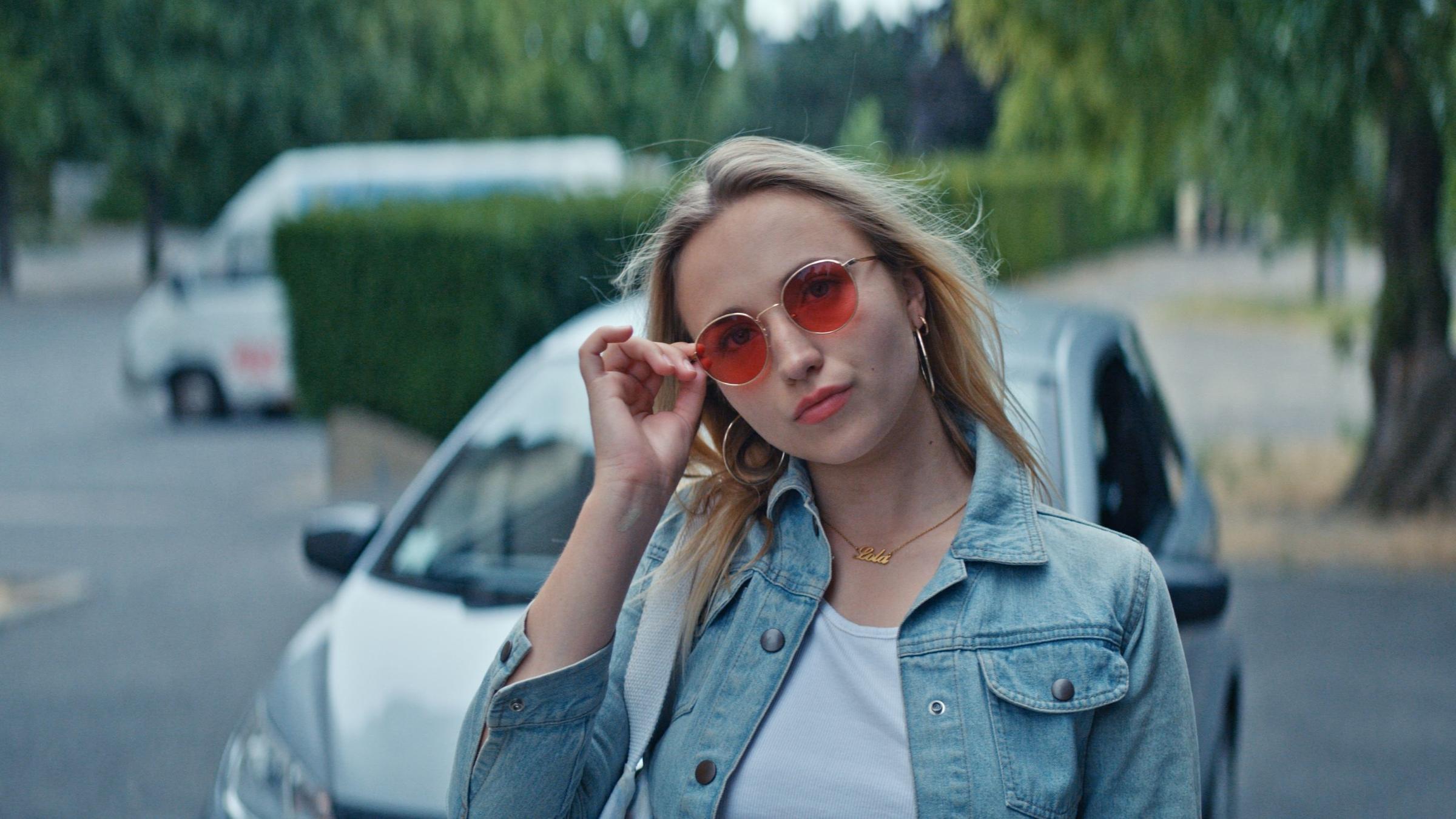 "Derby Girl - Lektionen": Auf einer Straße. Lola Bouvier (Chloé Jouannet) hält ihre rechte Hand an ihre farbige Sonnenbrille, neigt ihren Kopf ein wenig zur Seite und sie setzt einen coolen Gesichtsausdruck auf. Hinter ihr steht ein Auto.
