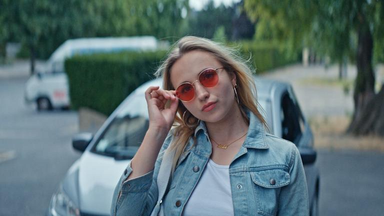 "Derby Girl - Lektionen": Auf einer Straße. Lola Bouvier (Chloé Jouannet) hält ihre rechte Hand an ihre farbige Sonnenbrille, neigt ihren Kopf ein wenig zur Seite und sie setzt einen coolen Gesichtsausdruck auf. Hinter ihr steht ein Auto.