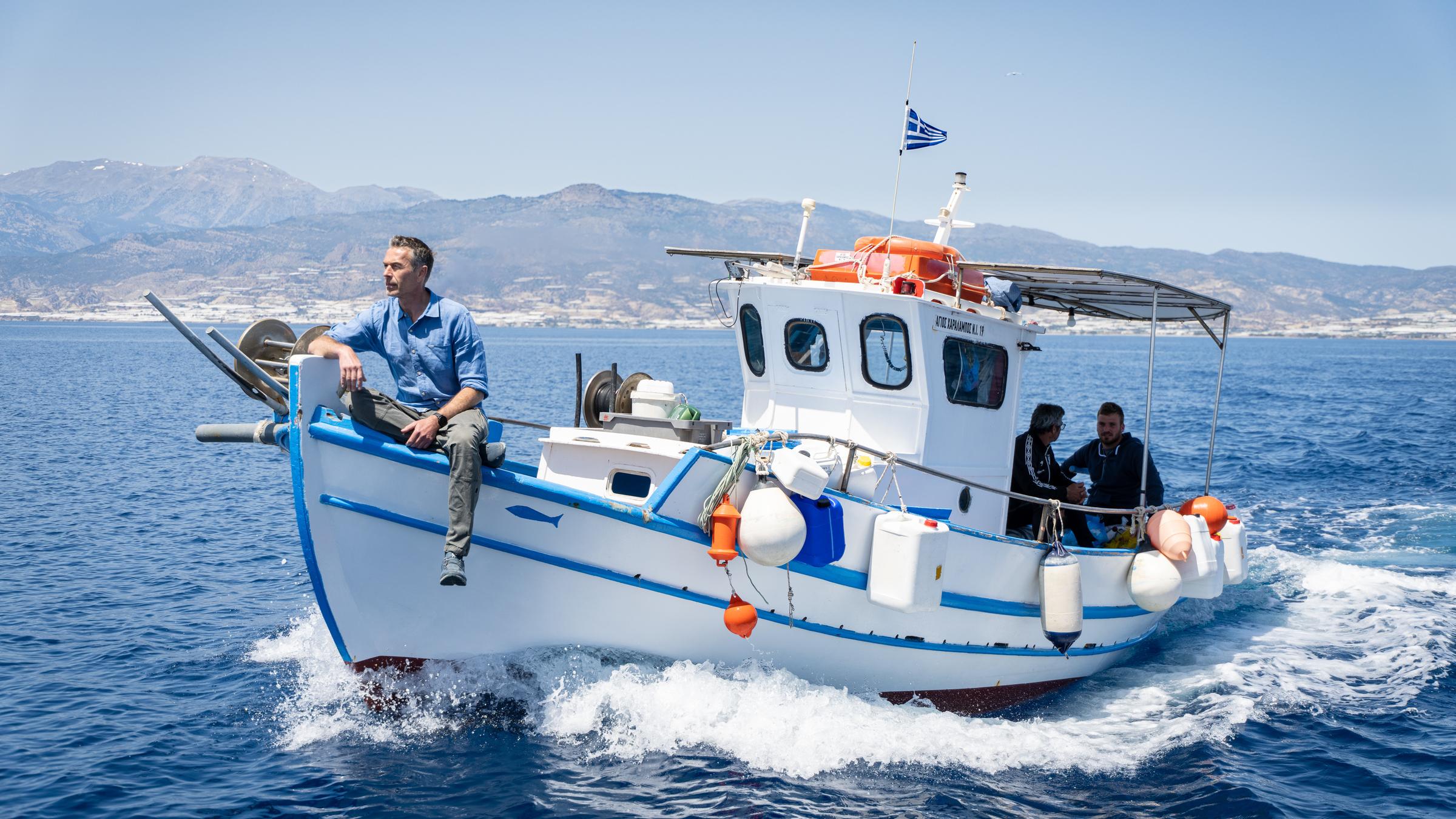 "Terra X: Rätselhafte Phänomene - Powerschnecken und haariges Eis": Moderator Dirk Steffens sitzt auf einem Fischerboot und blickt in Fahrtrichtung auf das Meer vor der Insel Chrysi. Mit an Bord sind zwei weitere Männer.