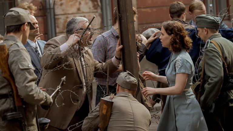 "ZDFzeit: Ein Tag im August - Mauerbau '61": Menschen, getrennt durch Stacheldraht, darunter eine junge Frau, Ingrid Taegner (Sandija Dovgāne), zwischen Grenzsoldaten, die einen älteren Mann auf der anderen Seite des provisorischen Zauns anschaut.