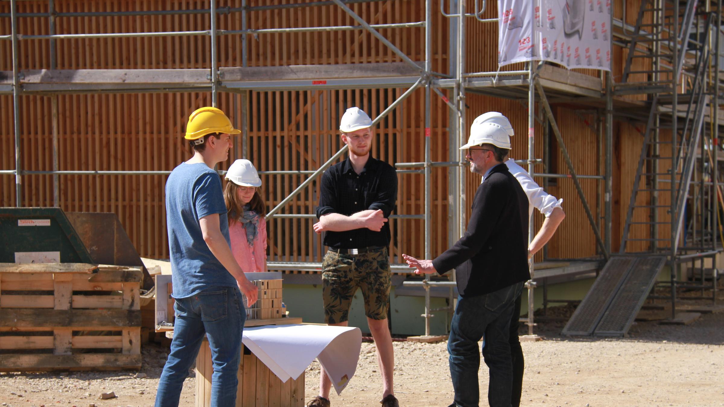 "plan b: Vier Wände für alle! - Wie Wohnraum bezahlbar werden kann": Vier Männer und eine Frau mit Bauhelmen stehen vor einem Rohbau mit Stahlgerüst und einem Modell und Bauplan auf einer Holzkabeltrommel und unterhalten sich.