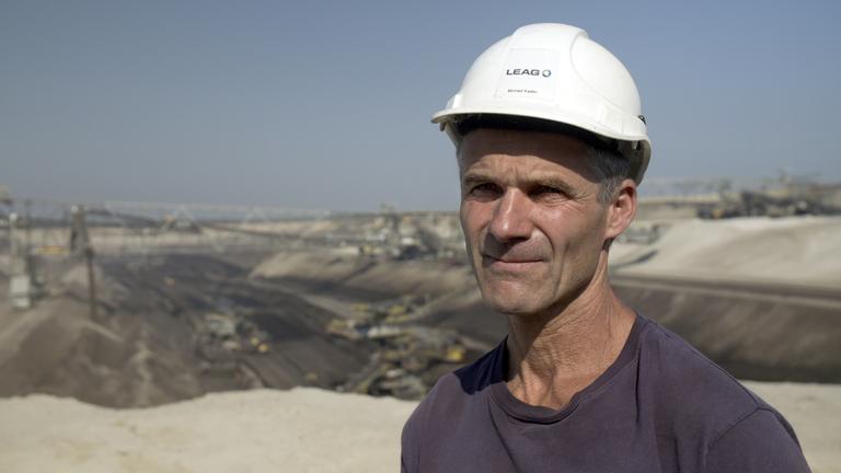 "37° Abgebaggert - Leben ohne Kohle": Ein Mann steht an der Kante zu einem Tagebau eines Kohlewerks. Er trägt einen weißen Bauhelm und schaut in die Kamera.