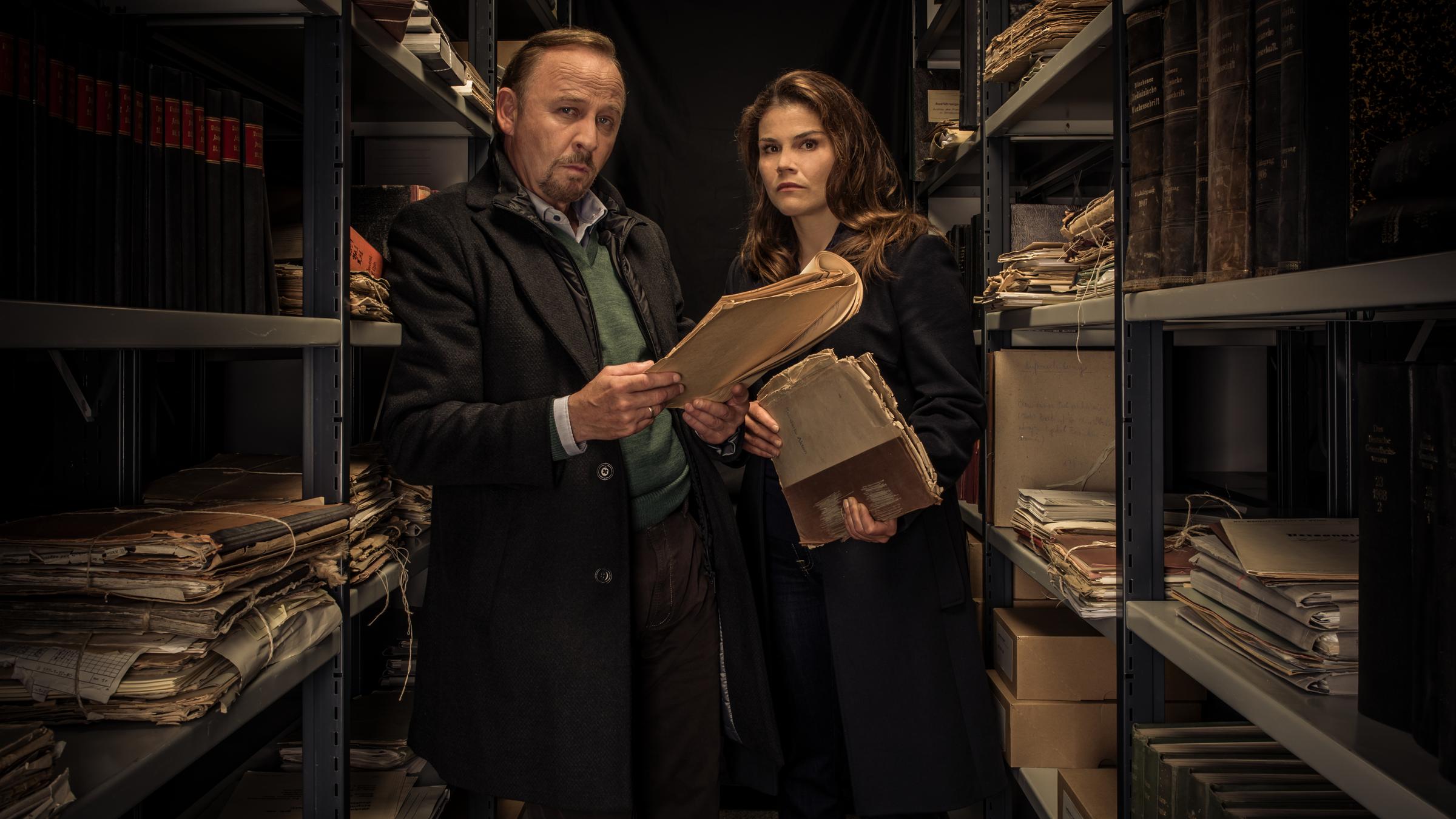 "Stralsund - Das Manifest": Nina Petersen (Katharina Wackernagel) und Karl Hidde (Alexander Held) stehen in einem düsteren Archiv und blicken mit Akten in den Händen in die Kamera.