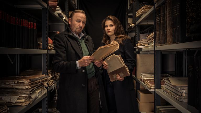 "Stralsund - Das Manifest": Nina Petersen (Katharina Wackernagel) und Karl Hidde (Alexander Held) stehen in einem düsteren Archiv und blicken mit Akten in den Händen in die Kamera.