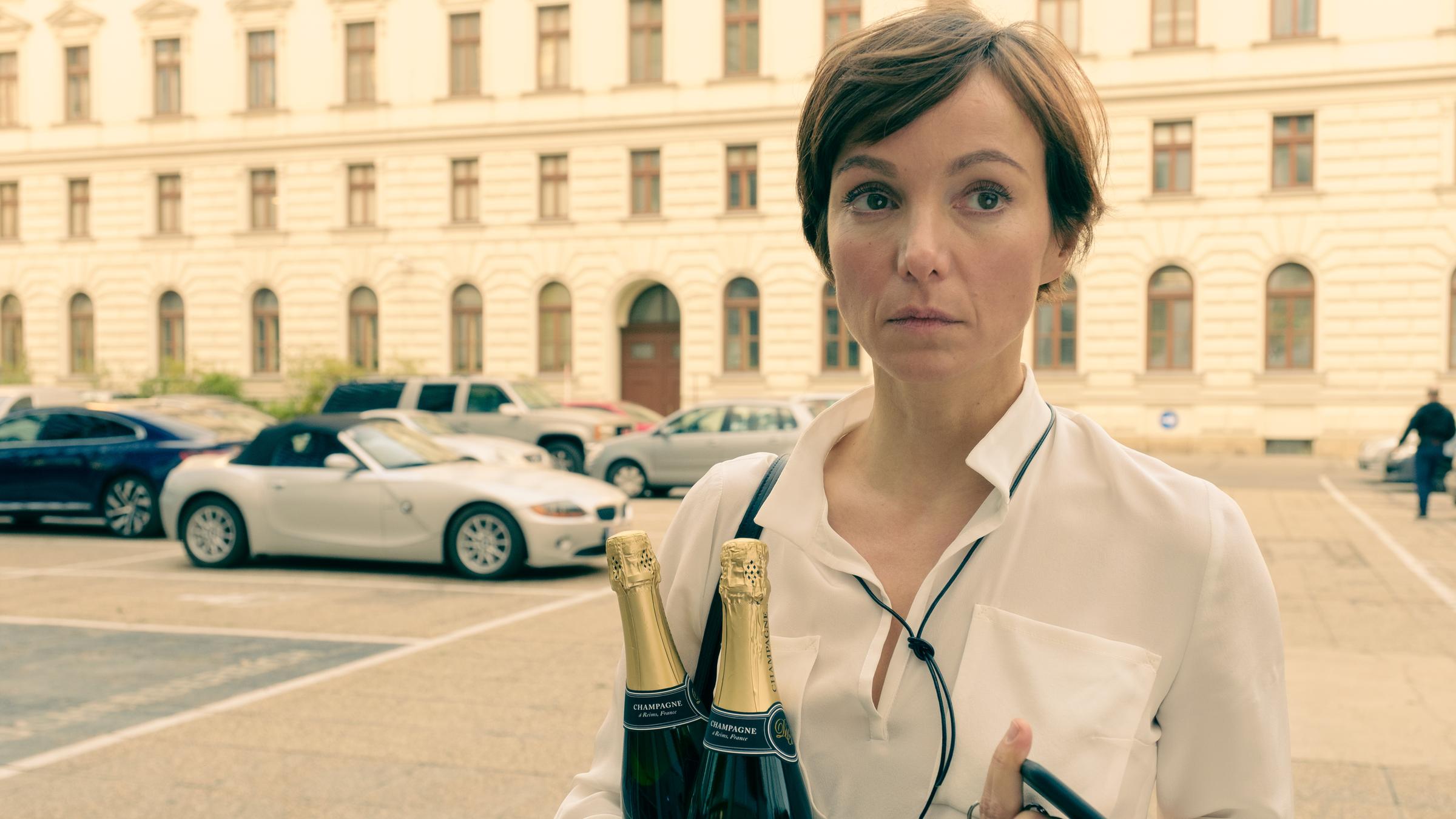 "Am Anschlag - Die Macht der Kränkung - MIRA": Mira (Julia Koschitz) blickt in die Ferne. In der linken Hand hält sie zwei Champagner Flaschen und in der rechten Hand ihr Mobiltelefon und einen Schlüsselbund. Im Hintergrund ist ein Gebäude zu sehen, vor dem Autos parken.