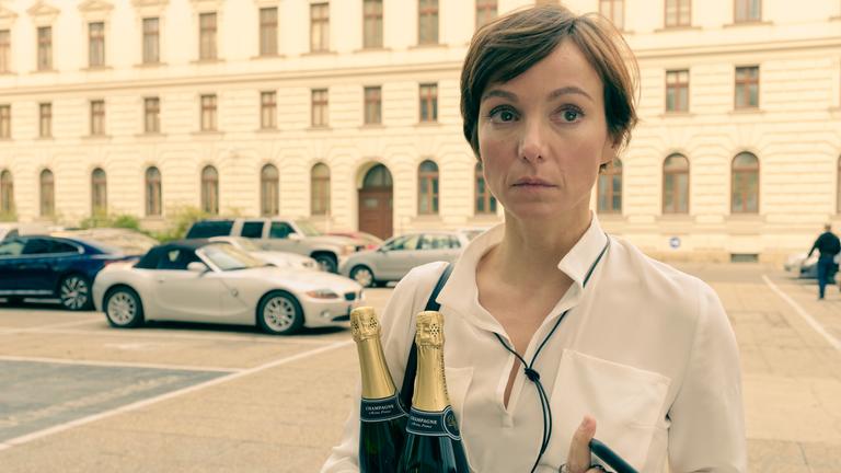 "Am Anschlag - Die Macht der Kränkung - MIRA": Mira (Julia Koschitz) blickt in die Ferne. In der linken Hand hält sie zwei Champagner Flaschen und in der rechten Hand ihr Mobiltelefon und einen Schlüsselbund. Im Hintergrund ist ein Gebäude zu sehen, vor dem Autos parken.