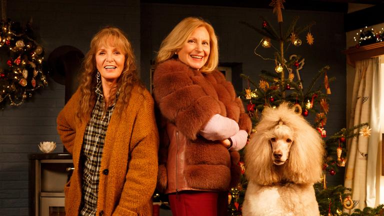 "Mona & Marie": Marie Decker (Ulrike Kriener) und ihre Schwester Mona Berthold (Maren Kroymann) stehen Rücken an Rücken in einem weihnachtlich geschmückten Wohnzimmer. Rechts von ihnen der weiße Königspudel Lucky.