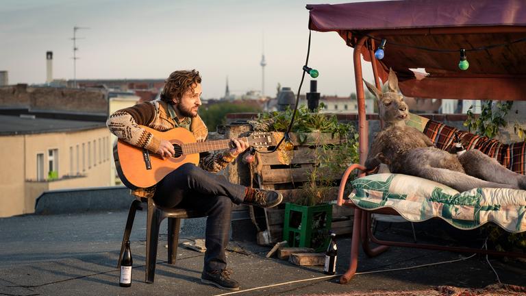 "Die Känguru-Chroniken": Marc-Uwe (Dimitrij Schaad) und das Känguru sitzen auf einer Berliner Dachterrasse. Marc-Uwe spielt Gitarre, während das Känguru es sich mit einem Bier auf einer Liege gemütlich gemacht hat.