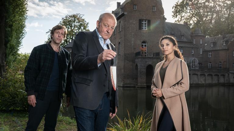 "Wilsberg - Einer von uns": In einem Park steht Wilsberg (Leonard Lansink) zwischen Ekki (Oliver Korittke) und Dr. Tessa Tilker (Patricia Meeden). In seiner rechten Hand hält Wilsberg ein blutiges Messer. Sie stehen vor einem Schlossgebäude.