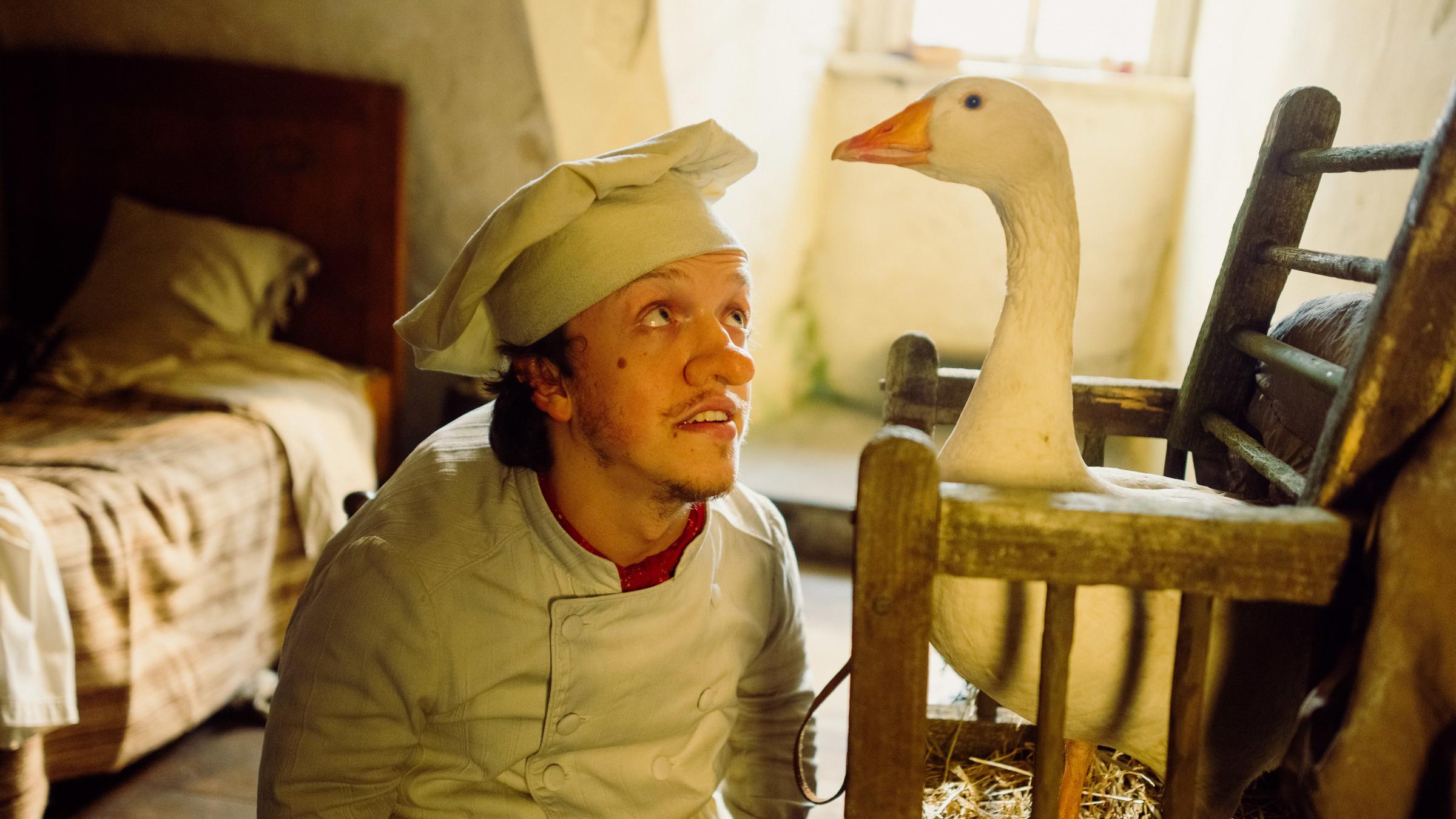 "Zwerg Nase": Inmitten seiner ärmlichen Kammer kniet liebevoll Zwerg Nase (Mick Mehnert) – mit übergroßer Nase, Kochmütze und in seinem weißen Arbeitskittel – vor einem Holzkäfig, aus dem keck die Gans Mimi hinausschaut.