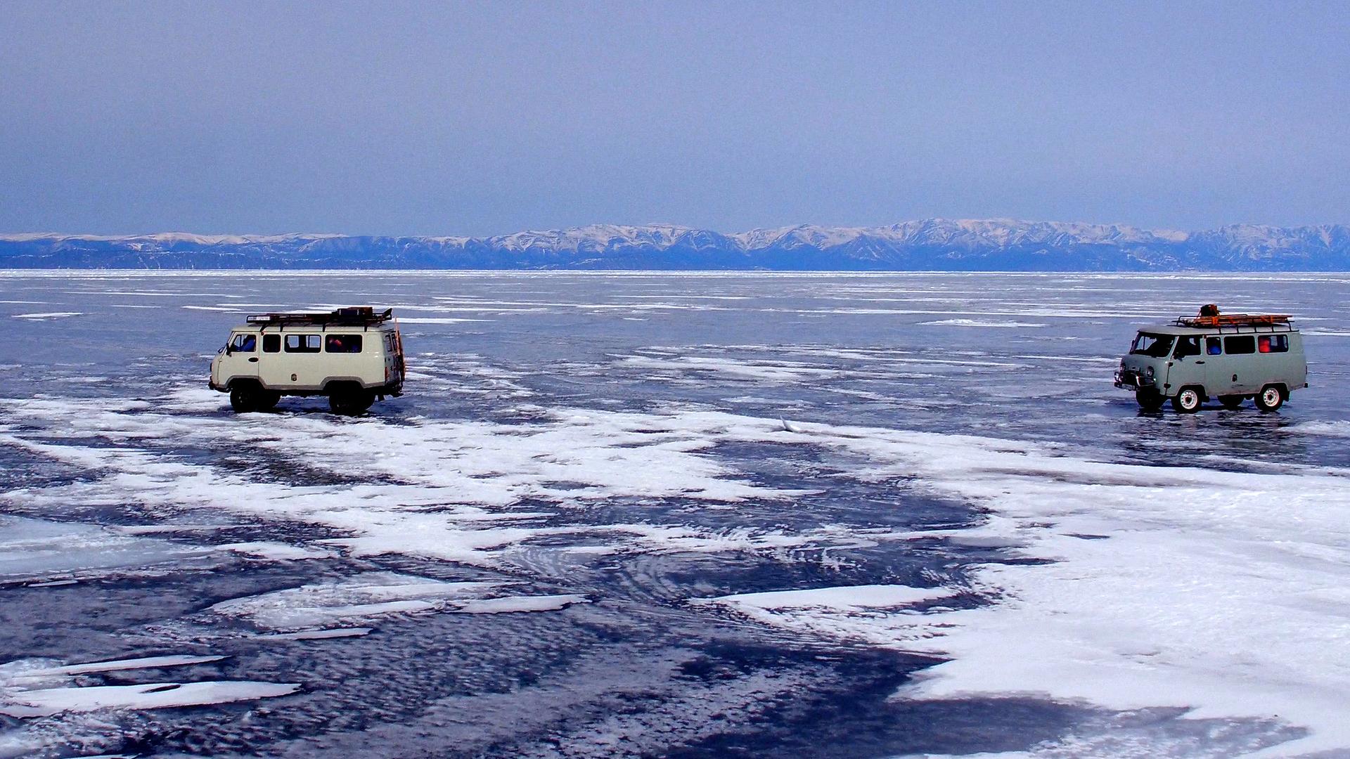 "Winterreise an den Baikalsee": Zwei Autos fahren über den tiefgefrorenen Baikalsee. Im Hintergrund schneebedeckte Berge und blauer Himmel.