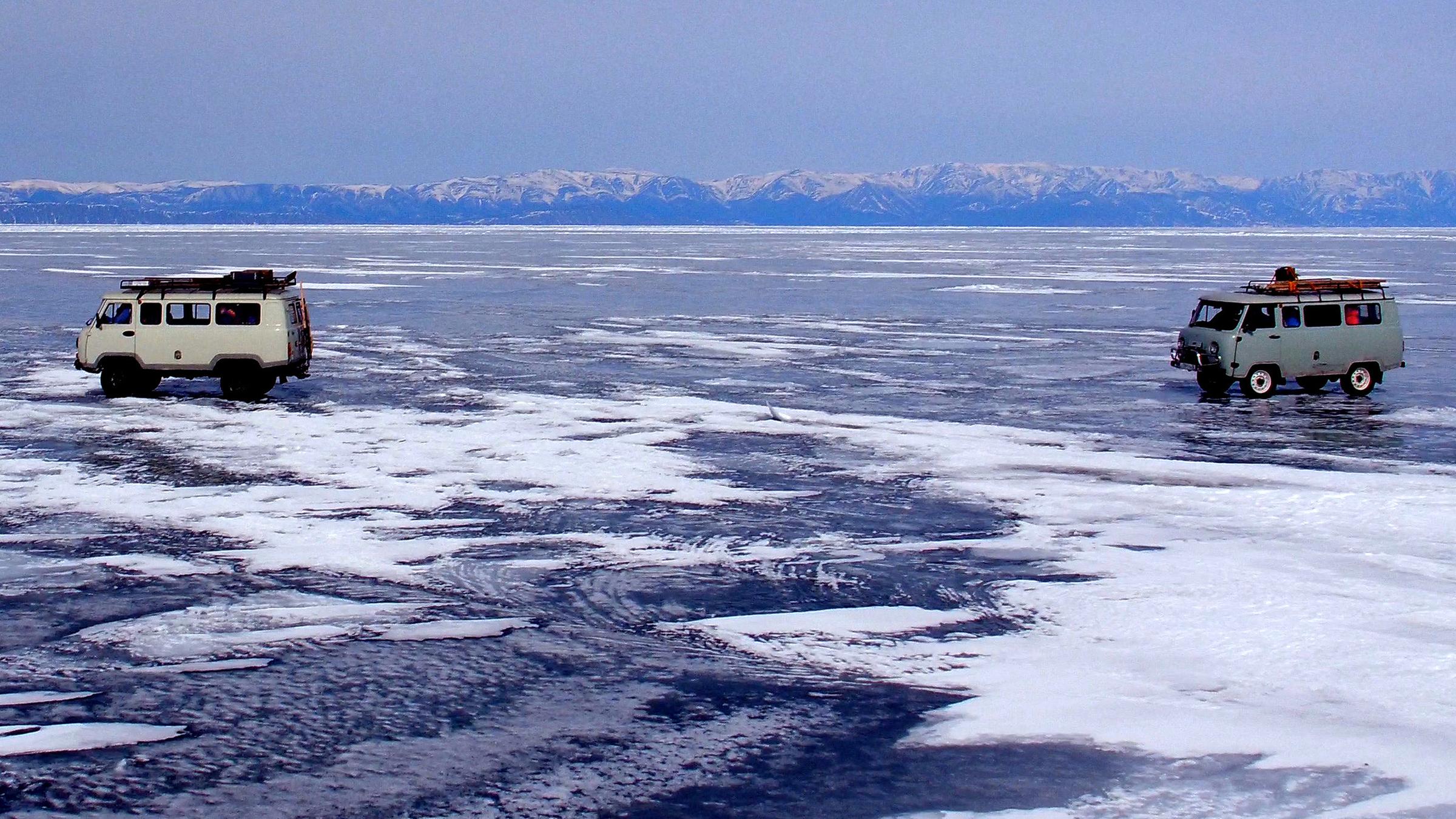 "Winterreise an den Baikalsee": Zwei Autos fahren über den tiefgefrorenen Baikalsee. Im Hintergrund schneebedeckte Berge und blauer Himmel.