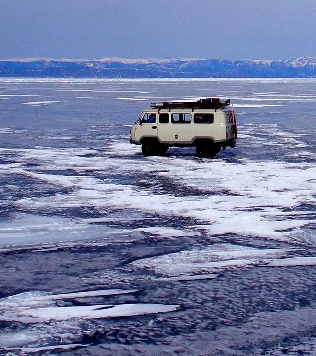 "Winterreise an den Baikalsee": Zwei Autos fahren über den tiefgefrorenen Baikalsee. Im Hintergrund schneebedeckte Berge und blauer Himmel.