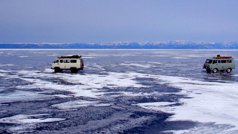 "Winterreise an den Baikalsee": Zwei Autos fahren über den tiefgefrorenen Baikalsee. Im Hintergrund schneebedeckte Berge und blauer Himmel.