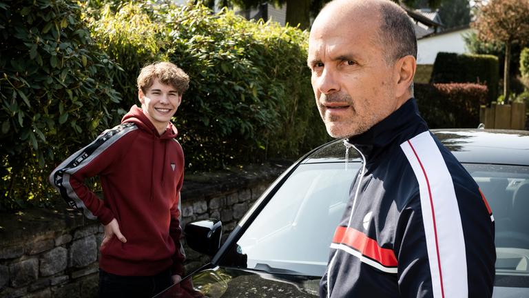 "Merz gegen Merz - Soraya": Draußen in der Einfahrt neben einem Auto: Leon Merz (Philip Noah Schwarz) steht lachend neben seinem Vater Erik Merz (Christoph Maria Herbst), der besorgt aus dem Bild blickt.