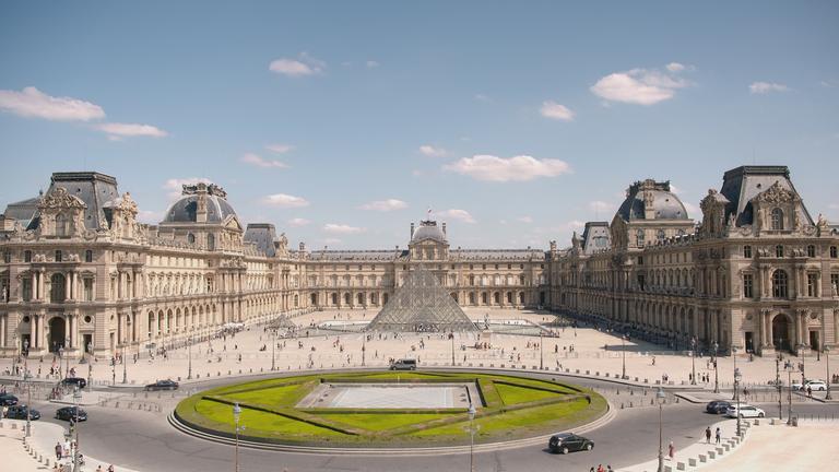 "Wunderwerke der Weltgeschichte: Der Louvre - Palast und Museum": Ein Bauwerk umrandet einen Platz. In der Mitte des Platzes ragt eine gläserne Pyramide aus der Erde: Der Louvre in Paris. Um eine kreisförmige Grasfläche führt eine Straße.