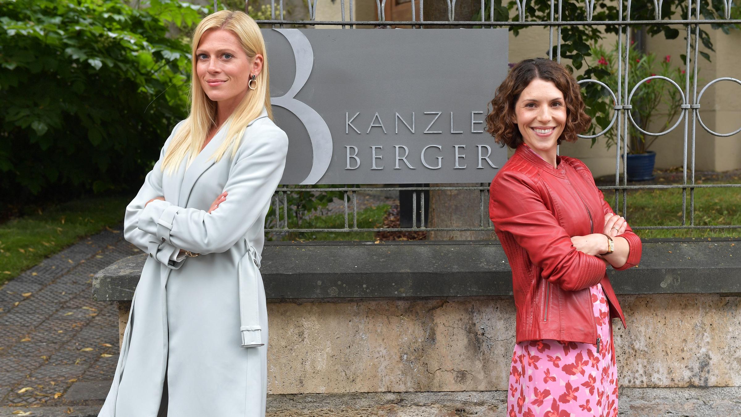 "Kanzlei Berger - Der Kollaps": Niki Berger (Nele Kiper) und ihre Schwester Caro (Eva Maria Reichert) stehen vor einer Mauer, auf der sich ein Metallzaun befindet. Daran ist ein dunkelgraues Schild mit dem Schriftzug "KANZLEI BERGER" befestigt. Die beiden haben sich den Rücken zugewandt und lächeln in die Kamera. Das Schild der Kanzlei ist zwischen ihnen zu sehen.