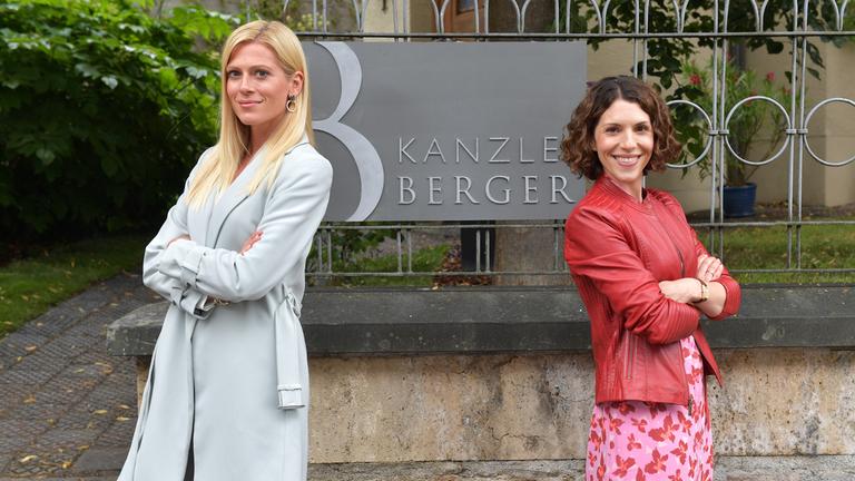 "Kanzlei Berger - Der Kollaps": Niki Berger (Nele Kiper) und ihre Schwester Caro (Eva Maria Reichert) stehen vor einer Mauer, auf der sich ein Metallzaun befindet. Daran ist ein dunkelgraues Schild mit dem Schriftzug "KANZLEI BERGER" befestigt. Die beiden haben sich den Rücken zugewandt und lächeln in die Kamera. Das Schild der Kanzlei ist zwischen ihnen zu sehen.