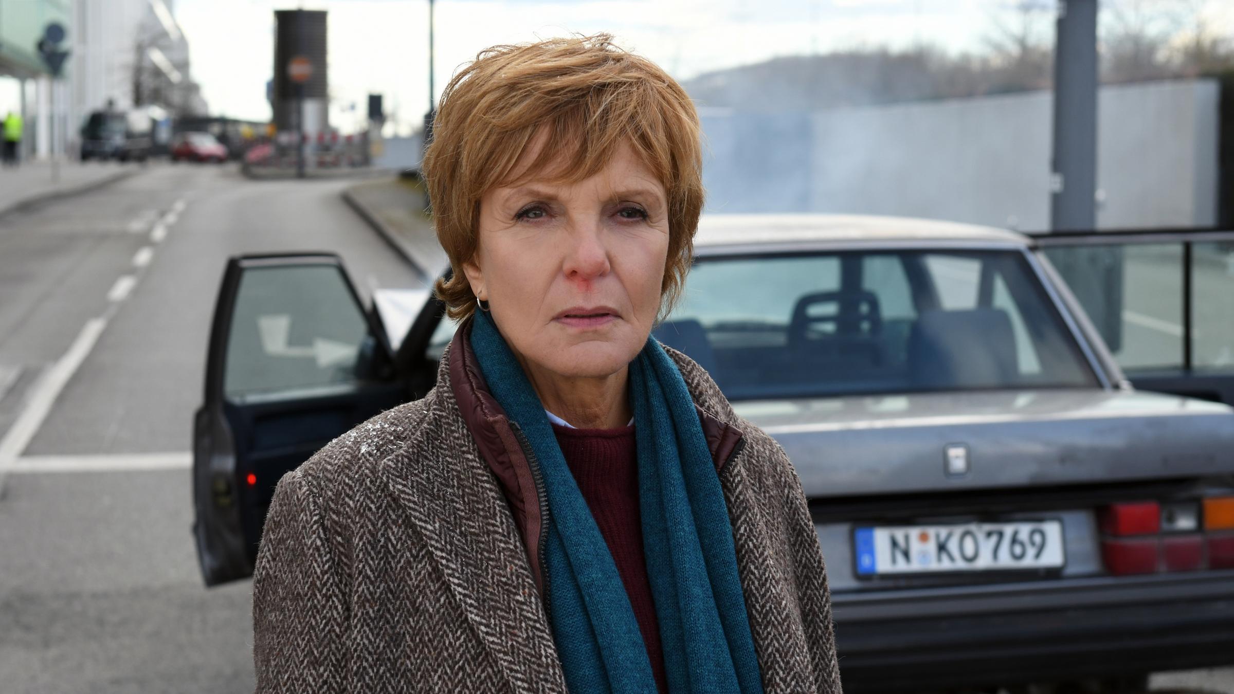 "Kommissarin Lucas - Nürnberg": Ellen Lucas (Ulrike Kriener) schaut nach vorne. Ihre Nase blutet. Im Hintergrund sieht man einen silbernen Pkw, bei dem beide Vordertüren geöffnet sind und Rauch von der Motorhaube aufsteigt.