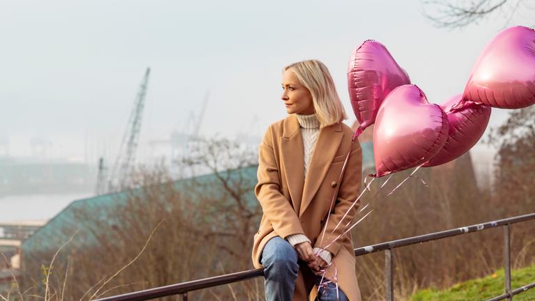 "Wie lange ist für immer?": Moderatorin Janin Ullmann sitzt auf einem Geländer, im Hintergrund ist der Hamburger Hafen zu sehen. Sie lässt den Blick in die Ferne schweifen. In den Händen hält sie die Fäden von vier Helium-Ballons in Herzform.