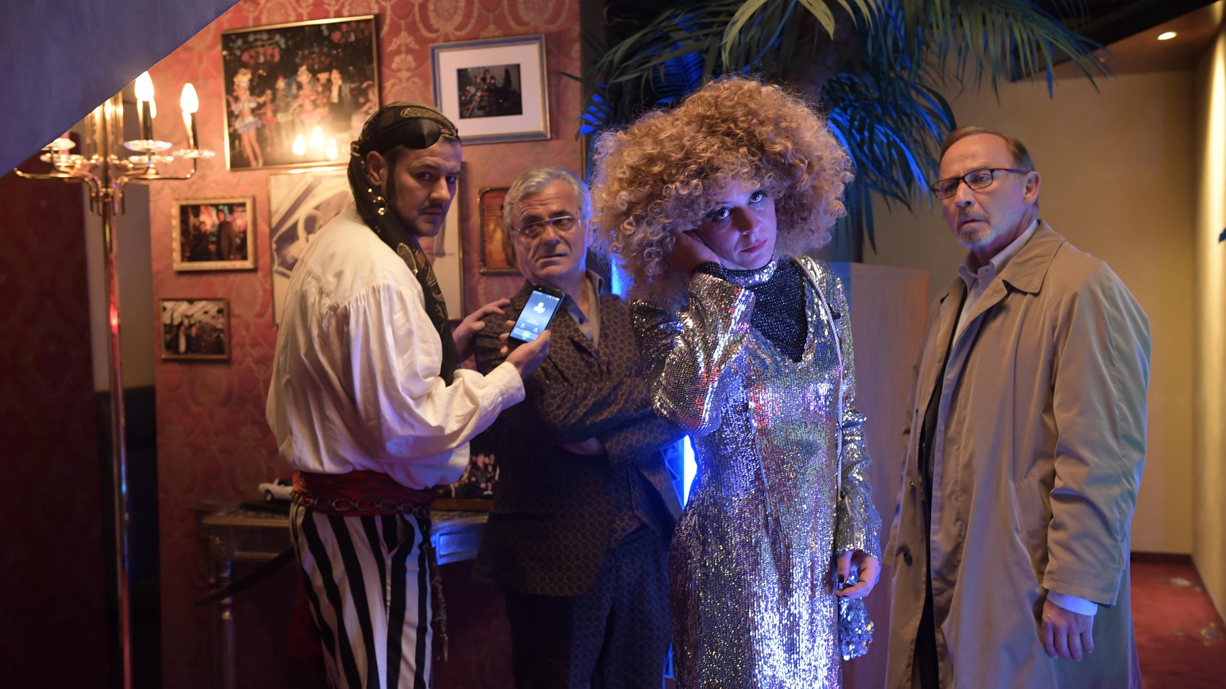 "München Mord - Der Letzte seiner Art": In einem bunt eingerichteten und spärlich beleuchteten Raum hält der als Pirat verkleidete Harald Neuhauser (Marcus Mittermeier) dem Arme verschränkenden Gustav Schmidinger (Martin Umbach) ein Handy vors Gesicht. Daneben hält sich Angelika Flierl (Bernadette Heerwagen) im Paillettenkleid die Hand ans Ohr und blickt mit dem neben stehendem Gustav Schmidinger (Martin Umbach) zielstrebig in die Kamera.