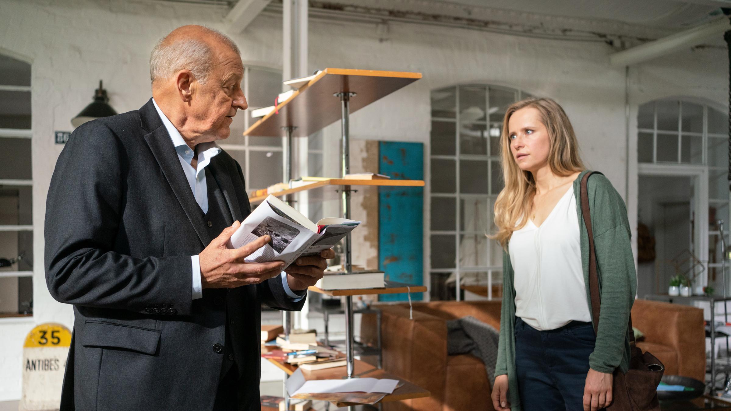 "Wilsberg - Überwachen und belohnen": Wilsberg (Leonard Lansink) und Christine Lau (Susanne Bormann) stehen in einem großen, hellen und wohnlich eingerichteten Loft. Wilsberg hält ein Buch  in den Händen. Hinter ihnen steht ein Bücherregal im Raum, aber die Bücher sind überall auf dem Boden verteilt.