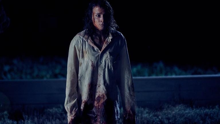 "Unbroken - Kapitel 1": Alex (Aylin Tezel) steht im Dunkeln mit starrem Blick auf einem Feld. Sie trägt ein helles, großes Hemd. In ihrem Gesicht und auf dem Hemd befindet sich Blut und Schmutz.