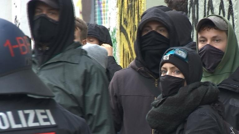 "Brandanschläge und Morddrohungen": Einige linke Demonstranten mit Mund-Nasen-Bedeckung im Gesicht stehen vor einer Hauswand neben einem Polizisten.