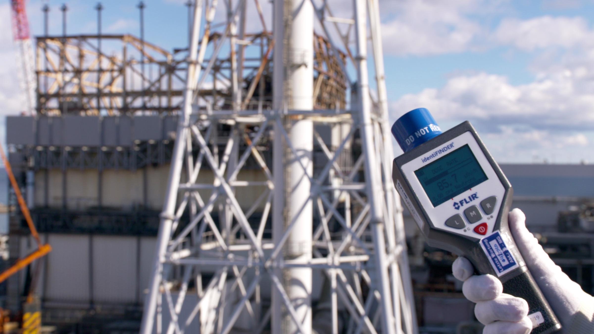 "Das Fukushima-Drama: Tsunami, Störfall, Langzeitfolgen": Handmessgerät mit Anzeige: "85,7 Mikrosievert pro Stunde": Im Hintergrund ist der zerstörte Block 1 von Fukushima Daiichi zu sehen.