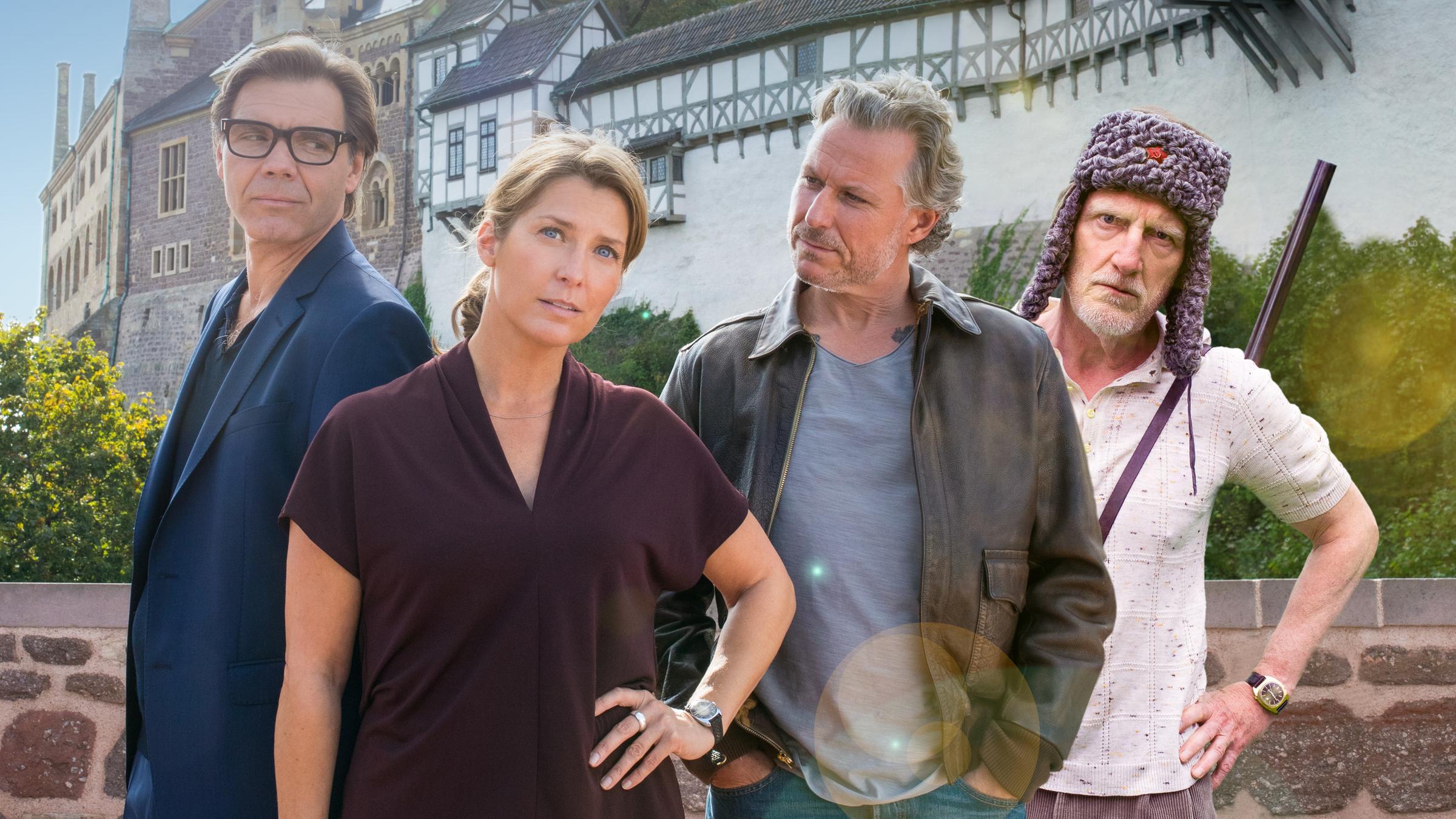 "Nächste Ausfahrt Glück - Juris Rückkehr": Katharina Wegener (Valerie Niehaus) steht mit etwas angestrengtem Gesichtsausdruck vor dem Hintergrund der Eisenacher Wartburg und hat eine Hand in die Hüfte gestützt. Links hinter ihr steht ihr Mann Georg (Max Hopp) und schaut sie misstrauisch an. Rechts neben ihr steht Juri Hoffmann (Dirk Borchardt) und betrachtet Katharina herausfordernd. Ganz rechts in der Gruppe steht Juris Vater Willi (Ernst Stötzner), mit einer russischen Fellmütze auf dem Kopf und einem Gewehr über die Schulter gehängt, und schaut dem Betrachter herausfordernd entgegen.