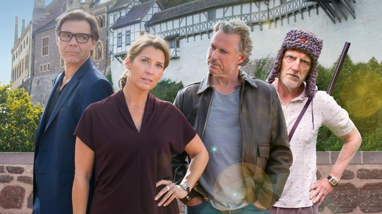 "Nächste Ausfahrt Glück - Juris Rückkehr": Katharina Wegener (Valerie Niehaus) steht mit etwas angestrengtem Gesichtsausdruck vor dem Hintergrund der Eisenacher Wartburg und hat eine Hand in die Hüfte gestützt. Links hinter ihr steht ihr Mann Georg (Max Hopp) und schaut sie misstrauisch an. Rechts neben ihr steht Juri Hoffmann (Dirk Borchardt) und betrachtet Katharina herausfordernd. Ganz rechts in der Gruppe steht Juris Vater Willi (Ernst Stötzner), mit einer russischen Fellmütze auf dem Kopf und einem Gewehr über die Schulter gehängt, und schaut dem Betrachter herausfordernd entgegen.