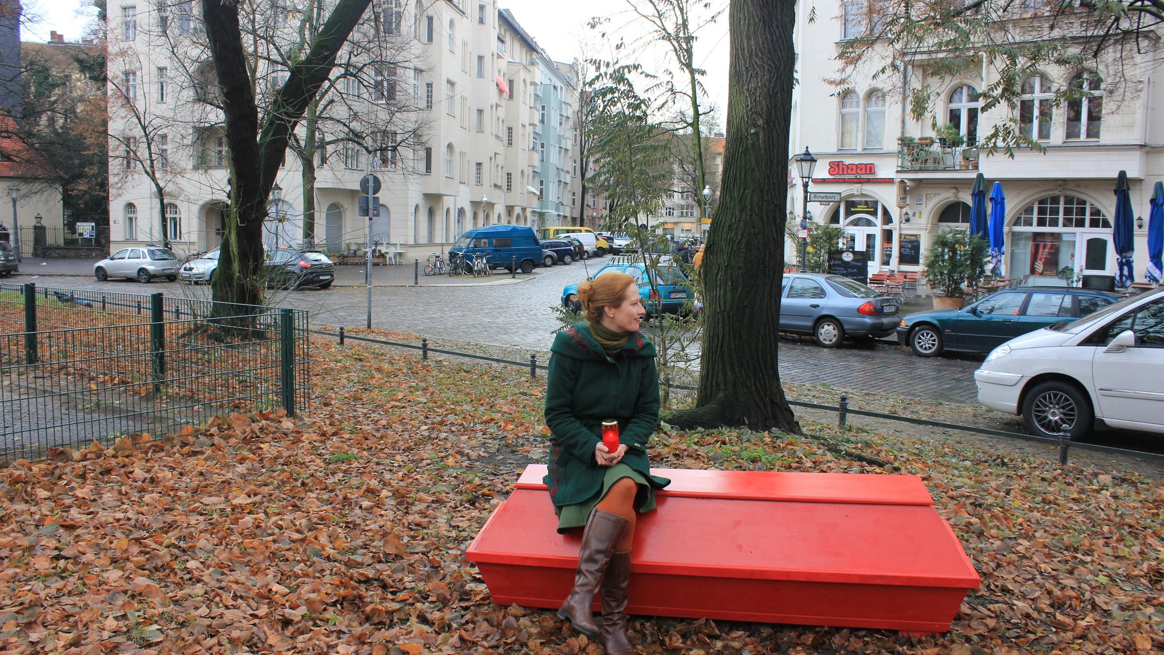 "ZDFzoom: Das Virus und seine Opfer - Wie Corona das Sterben verändert": Die Bestatterin Sarah Benz sitzt auf einem roten Sarg in einem mit Laub bedeckten Park. Im Hintergrund sieht man eine Nebenstraße mit Häusern und parkenden Autos.
