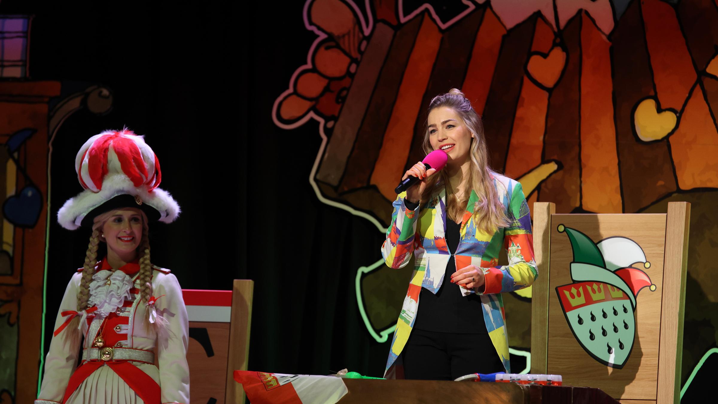 "Kölle Alaaf - Die Mädchensitzung": Sitzungspräsidentin Martina Kratz, moderiert stehend, mittig im Elferratsgestühl, mit einem Mikrofon in der Hand. Sie trägt einen Frack mit bunten Flecken.