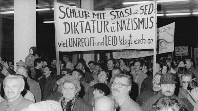 "Wendezeit - Sturm auf die Stasi-Zentrale": Schwarz-Weiß-Aufnahme: Eine Gruppe von Menschen in einem Gebäude. Einige halten Plakat hoch.