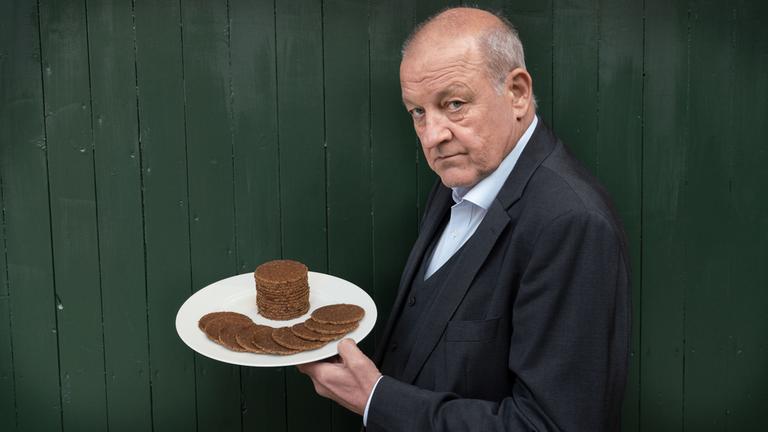 "Wilsberg: Unser tägliches Brot": Wilsberg (Leonard Lansink) steht vor einer Bretterwand und hält einen Teller mit dunklen runden Scheiben Pumpernickelbrot hoch. Er schaut in die Kamera.