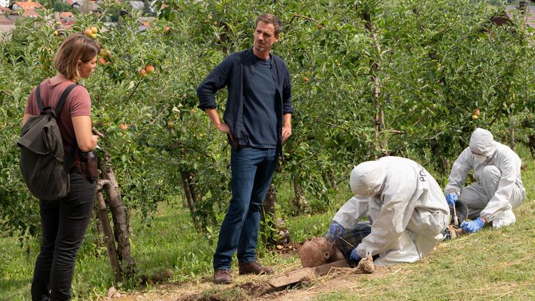 "Waldgericht - Ein Schwarzwaldkrimi - Die schwarze Frau": Auf einer Streuobstwiese liegt ein Toter. Die Kommissare Maris Bächle (Jessica Schwarz) und Konrad Diener (Max von Thun) stehen daneben und besprechen sich. Neben dem Toten knien zwei Spurensicherer.