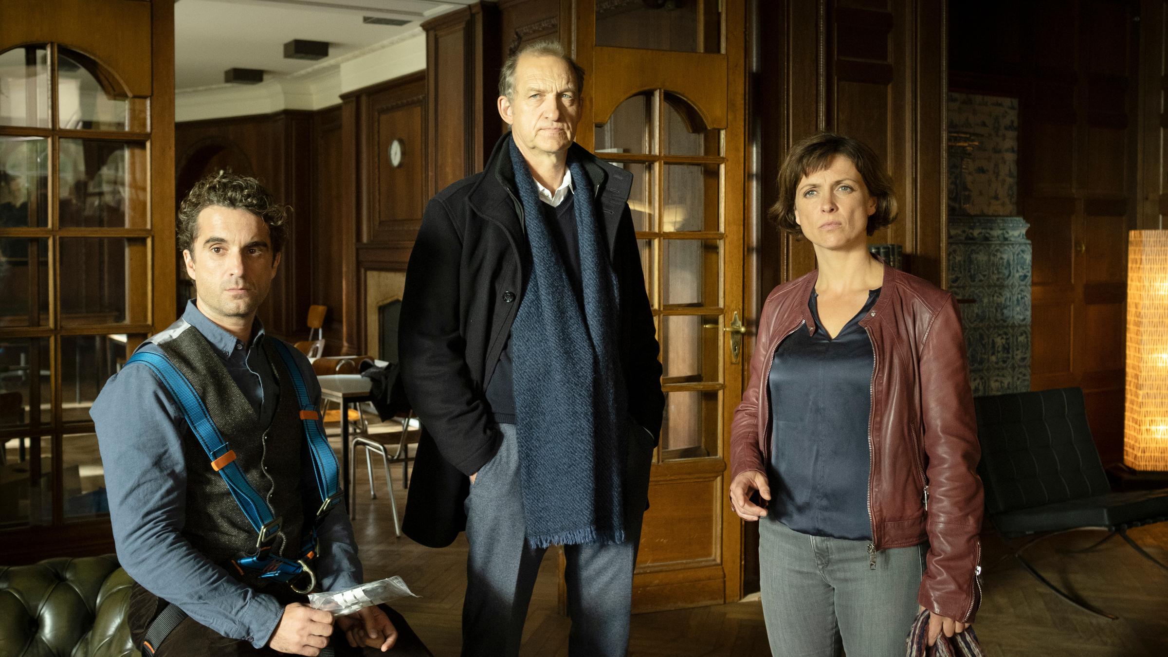 "Nord Nord Mord – Sievers und der goldene Fisch": Ina Behrendsen (Julia Brendler), Carl Sievers (Peter Heinrich Brix) und Hinnerk Feldmann (Oliver Wnuk) befinden sich in einem mit Eichenholz getäfelten Empfangsraum in der Schule. Im Hintergrund sind eine geöffnete Tür mit Glasscheiben, eine Uhr, ein Kamin und Tische mit Stühlen zu sehen.