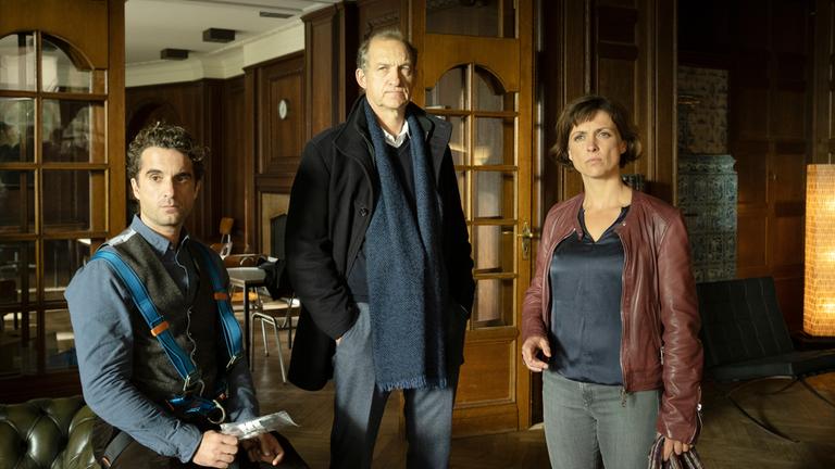 "Nord Nord Mord – Sievers und der goldene Fisch": Ina Behrendsen (Julia Brendler), Carl Sievers (Peter Heinrich Brix) und Hinnerk Feldmann (Oliver Wnuk) befinden sich in einem mit Eichenholz getäfelten Empfangsraum in der Schule. Im Hintergrund sind eine geöffnete Tür mit Glasscheiben, eine Uhr, ein Kamin und Tische mit Stühlen zu sehen.