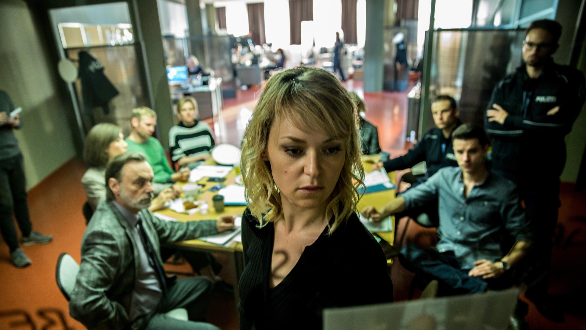 "Kommissarin Heller - Panik": Winnie Heller (Lisa Wagner) steht vor einer Glaswand und schaut sich ein Bild an. Im Hintergrund sitzen ihre Kollegen an einem runden Tisch.