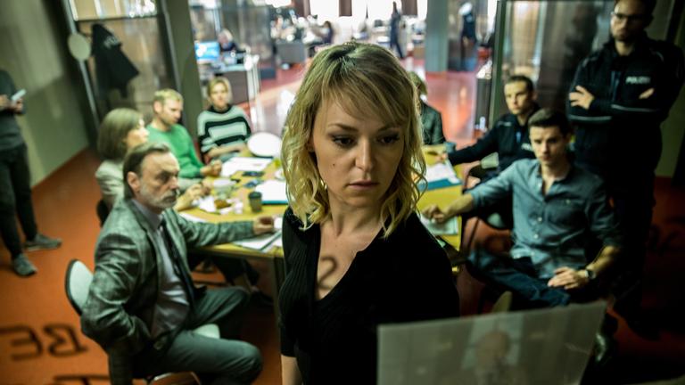 "Kommissarin Heller - Panik": Winnie Heller (Lisa Wagner) steht vor einer Glaswand und schaut sich ein Bild an. Im Hintergrund sitzen ihre Kollegen an einem runden Tisch.