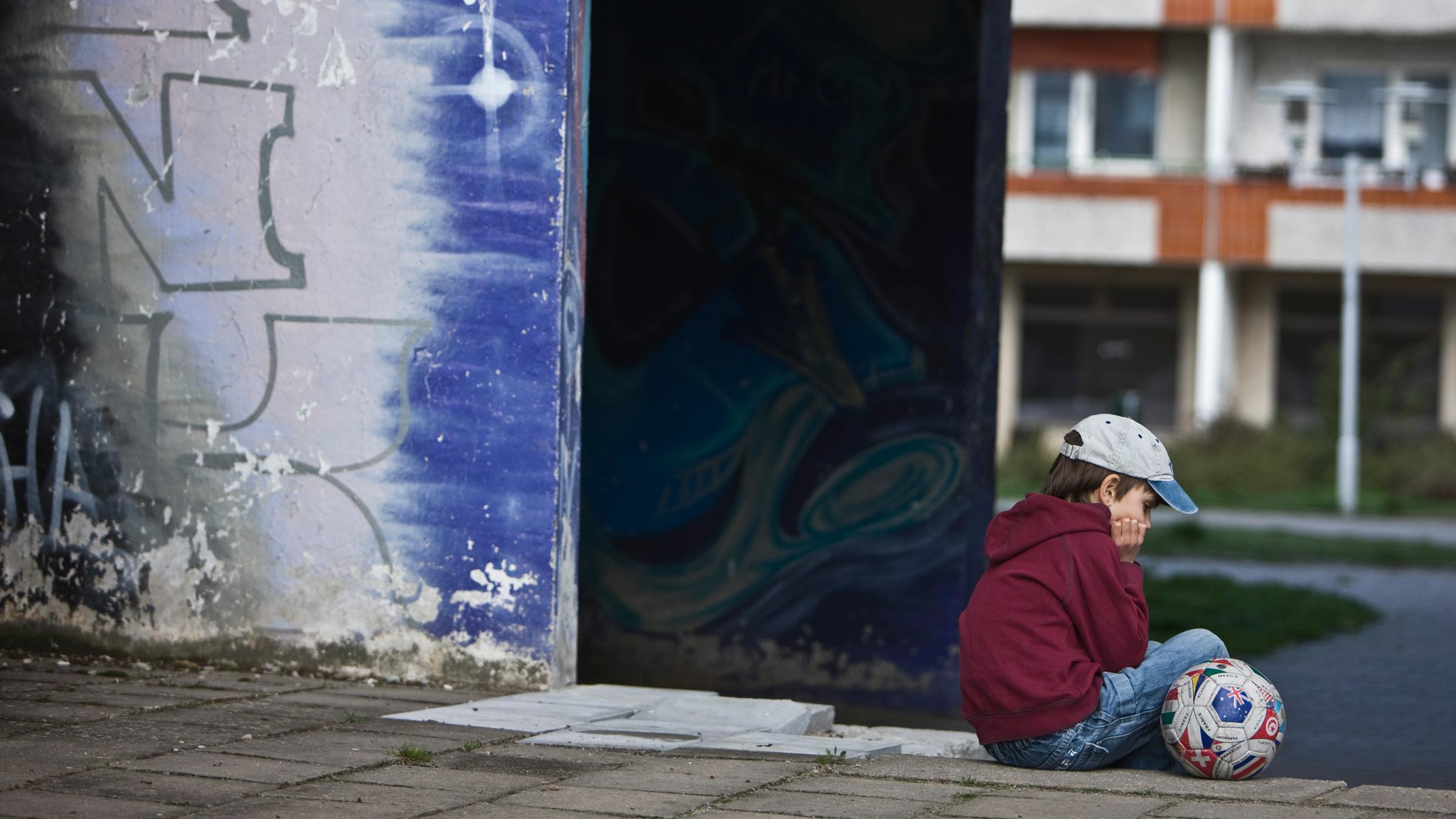 "ZDF.reportage: Alltag Kinderarmut -  Kein Geld - keine Chance?": Ein Junge seitlich zur Kamera, am rechten Bildrand mit Jeans, einer Kappe und einem roten Sweater. Er sitzt alleine mit seinem Fußball in einem ärmlichen Wohnviertel.
